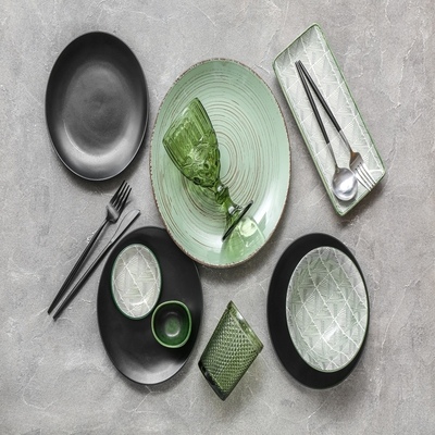 Mesa posta vista de cima com pratos pretos e verdes, travessas de vidro texturizado, talheres pretos e acessórios decorativos em estilo contemporâneo.