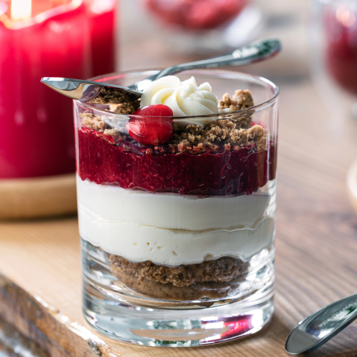 Taça de sobremesa com camadas de creme branco, compota de frutas vermelhas e farofa crocante, decorada com chantilly e uma cereja, servida com colher.