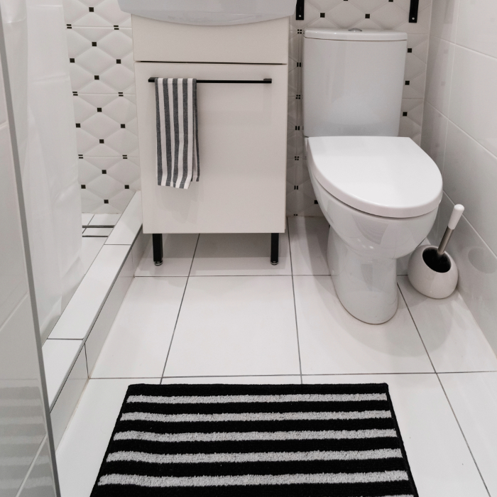Banheiro branco com tapete listrado preto e branco diante da pia e vaso sanitário, em ambiente compacto e moderno.