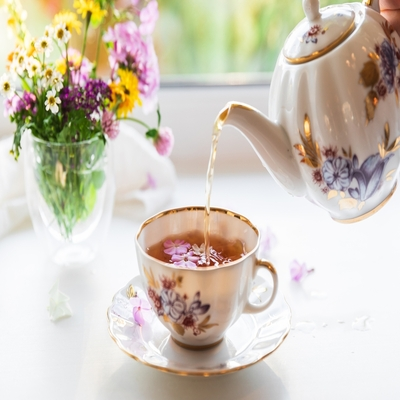 Xícara de porcelana estampada com flores e detalhes dourados recebendo chá, acompanhada de bule e arranjo floral em um ambiente iluminado.