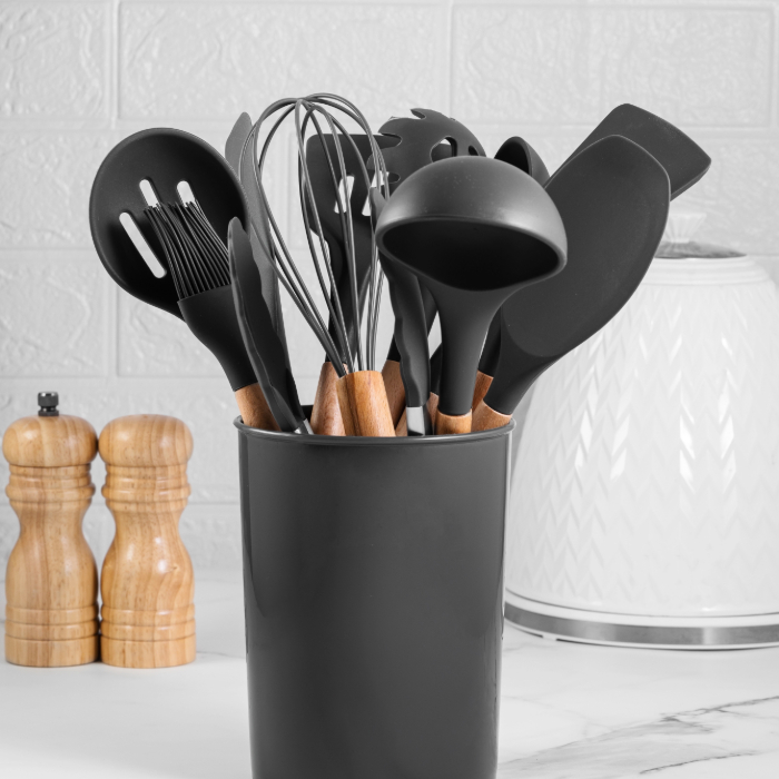 Conjunto de utensílios de cozinha com cabos de madeira e cabeças de silicone preto, organizados em um suporte preto sobre uma bancada branca, com moedores de madeira ao fundo.