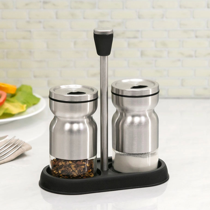 Conjunto de saleiro e pimenteiro em inox com base e suporte preto, posicionado sobre uma mesa com utensílios ao fundo.
