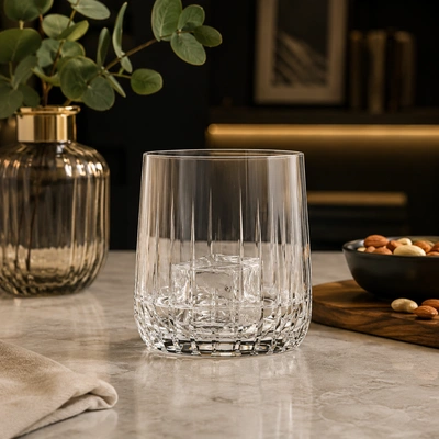 Copo de vidro lapidado com cubos de gelo sobre bancada de pedra, decorado com vaso, petiscos e iluminação aconchegante ao fundo.