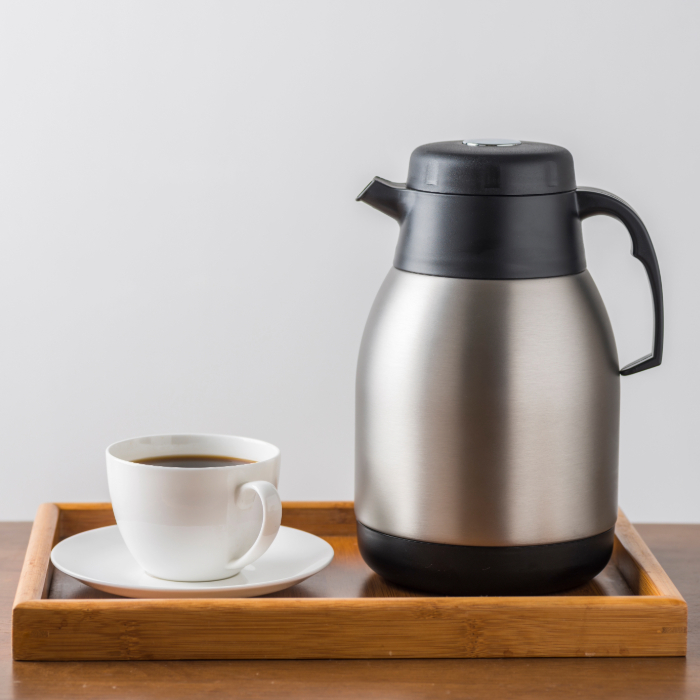 Garrafa térmica de inox com alça preta ao lado de uma xícara de café sobre bandeja de madeira.