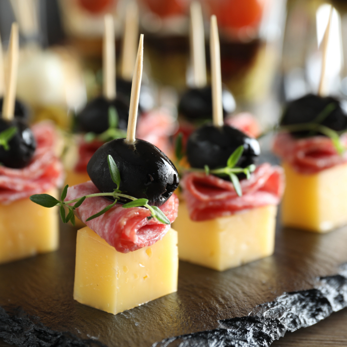 Petiscos gourmet com cubos de queijo, fatias de salame, azeitonas pretas e ramos de ervas frescas, espetados com palitos de dente sobre uma tábua de ardósia.