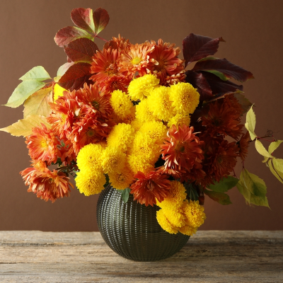 Adorno decorativo em formato de vaso com flores amarelas e alaranjadas, disposto sobre superfície de madeira.