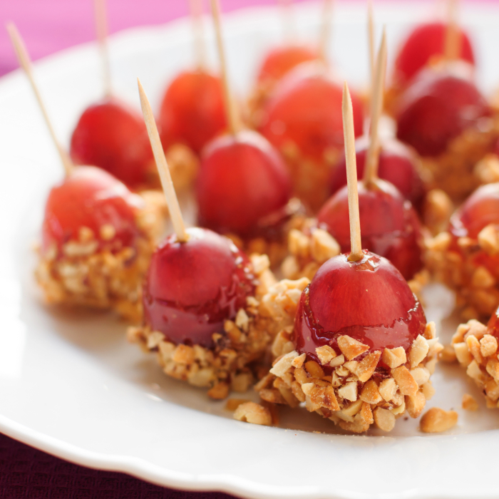 Uvas caramelizadas com cobertura de amendoim picado, espetadas com palitos de dente, dispostas em um prato branco.