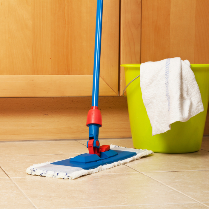 Mop azul e vermelho ao lado de balde verde com pano branco pendurado, sobre piso de cozinha.