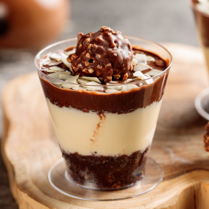 Sobremesa servida em copo transparente com camadas de creme branco e bolo de chocolate, coberta com ganache de chocolate, lâminas de amêndoas e finalizada com um bombom de chocolate crocante no topo.