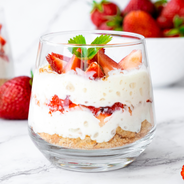 Sobremesa de morango servida em uma taça  de sobremesa, com camadas de creme branco, pedaços de morango fresco e farofa crocante na base, decorada com folhas de hortelã no topo. Morangos frescos ao fundo.
