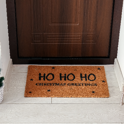 Capacho de porta marrom com mensagem natalina “HO HO HO”, posicionado em frente à porta de entrada.