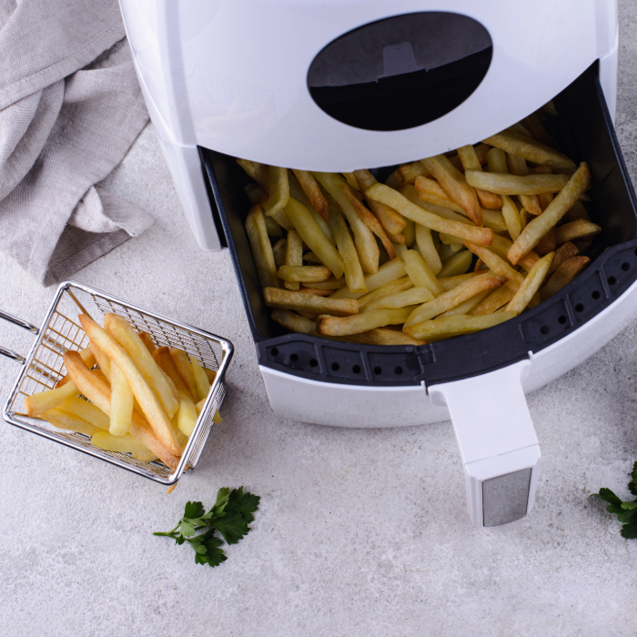 Fritadeira elétrica air fryer branca com gaveta aberta, cheia de batatas fritas douradas. Ao lado, pequena cesta metálica com batatas e ramos de salsa sobre uma superfície clara.