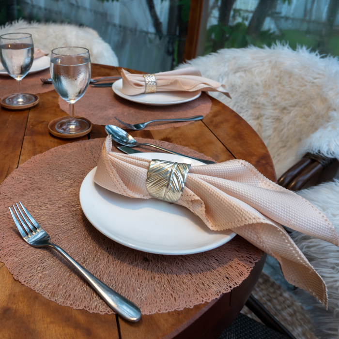 Mesa posta com guardanapos rosados presos por argolas prateadas com relevo, sobre sousplats rosé.