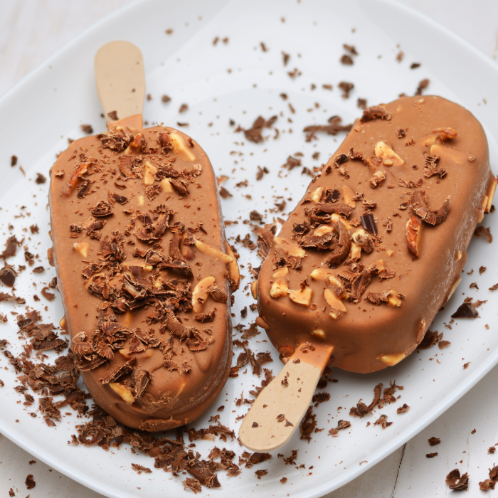 Sorvetes no palito cobertos com chocolate ao leite e pedaços de castanha, decorados com raspas de chocolate em um prato branco.