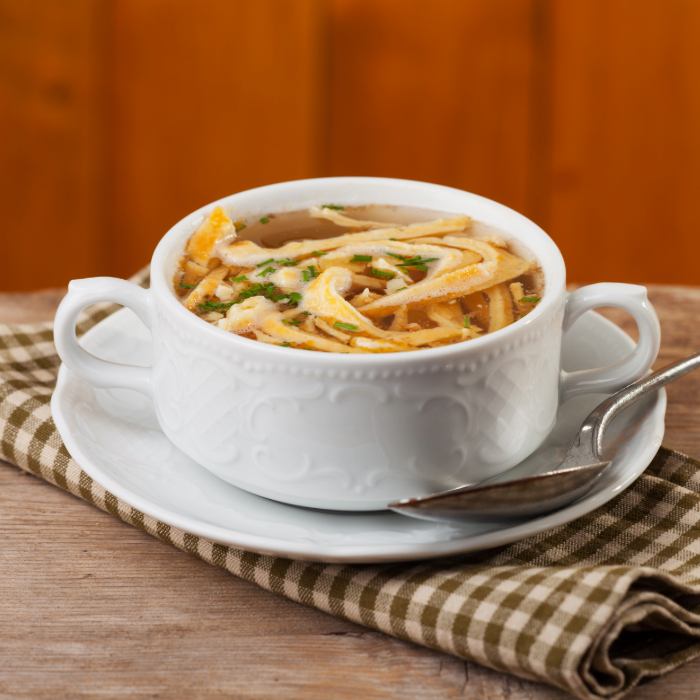 Sopa quente servida em uma consomê branca com alças, acompanhada de colher, em cima de um pires.