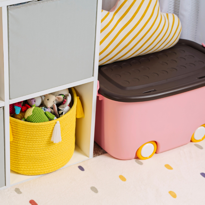 Cestos e caixas organizadoras em um ambiente infantil, incluindo um cesto de corda amarelo com brinquedos e uma caixa rosa com rodinhas ao lado de uma estante branca.