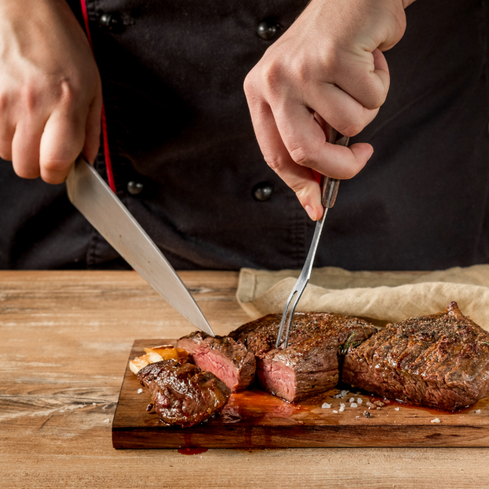 Chef fatiando um suculento corte de carne malpassada sobre uma tábua de madeira, usando faca e garfo trinchante.