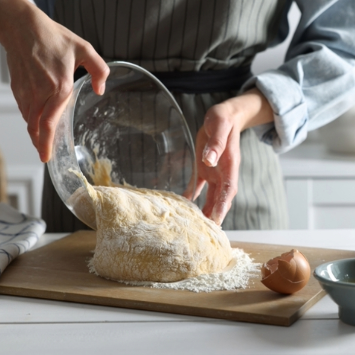 Pessoa despejando massa fermentada de um bowl de vidro sobre uma tábua enfarinhada, com uma casca de ovo ao lado.
