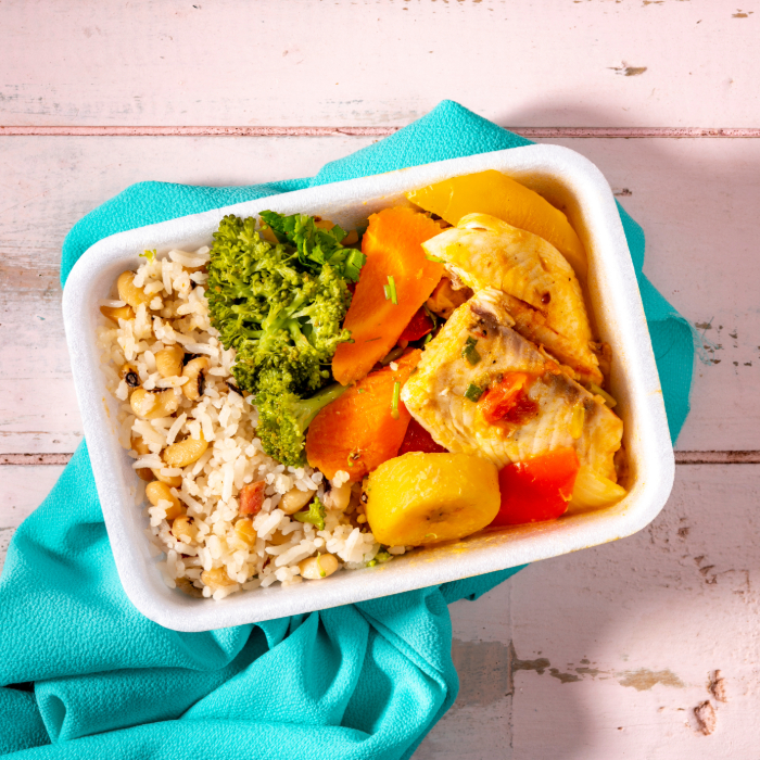 Marmita contendo arroz com feijão-fradinho, brócolis, cenoura, batata e peixe ensopado, apoiada sobre um pano azul em fundo de madeira clara.