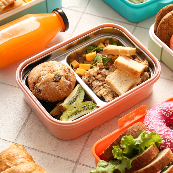 Lancheira com compartimentos contendo muffin de frutas, fatias de kiwi, salada com cubos de manga, aveia e torradas, acompanhada de garrafa de suco laranja em fundo claro.