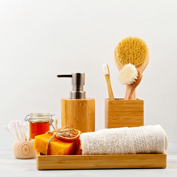 Conjunto de acessórios de banheiro com base de bambu, incluindo dispenser de sabão, escova corporal, sabonete em barra, toalha felpuda, escova de dente de bambu, potinho de mel, cotonetes e esponjas naturais, sobre superfície clara.