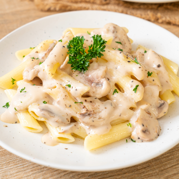 Prato de penne ao molho branco com pedaços de cogumelos, decorado com salsa fresca, servido em prato branco sobre mesa de madeira.