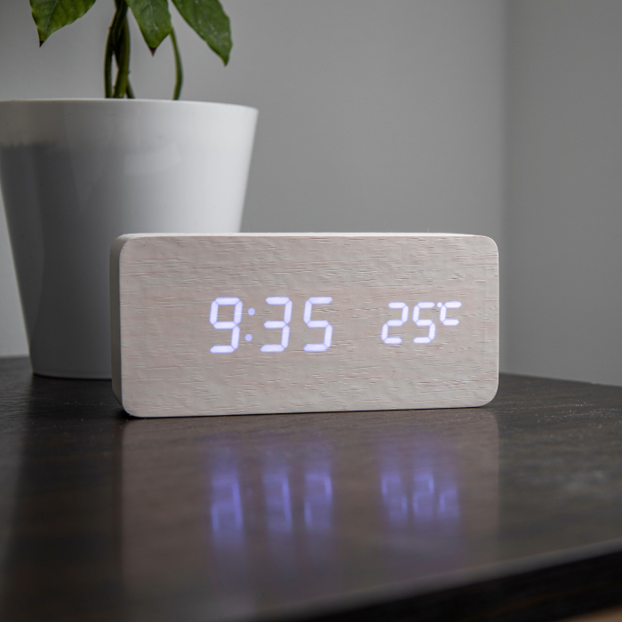 Relógio digital com estrutura em madeira clara mostrando hora e temperatura sobre mesa escura, ao lado de vaso branco com planta