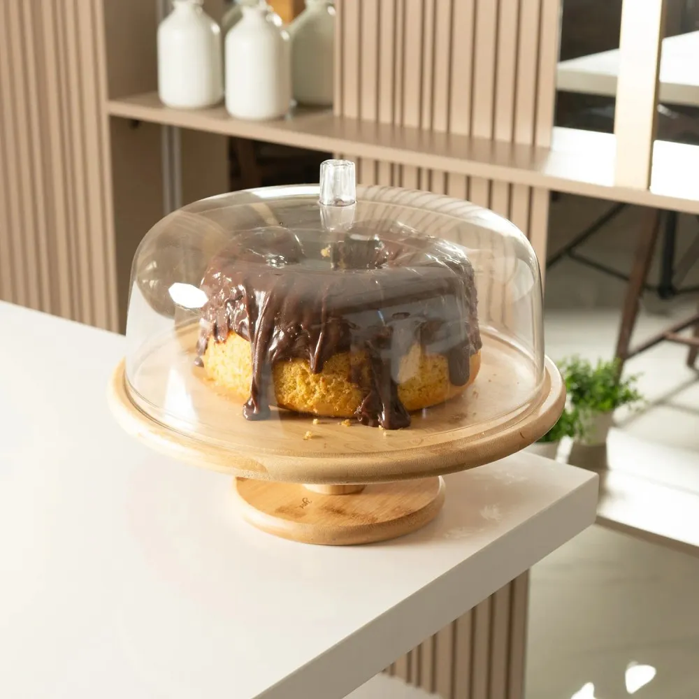 Boleira de madeira com pé elevado e tampa transparente com puxador, protegendo bolo caseiro com cobertura de chocolate sobre bancada branca.