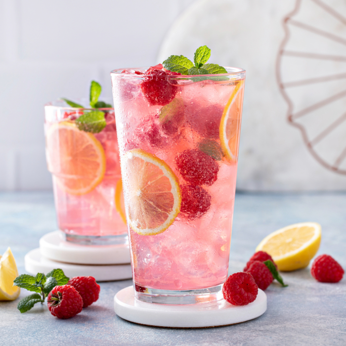 Duas bebidas refrescantes cor-de-rosa servidas em copos altos com gelo, fatias de limão, framboesas e folhas de hortelã, dispostas sobre porta-copos brancos em uma mesa azul, com limões e framboesas ao redor.