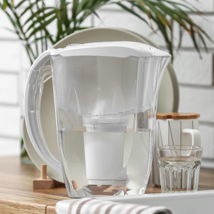Jarra de água transparente com filtro embutido e tampa branca, posicionada sobre uma mesa de madeira, com pratos, copo e utensílios ao fundo em um ambiente iluminado e moderno.