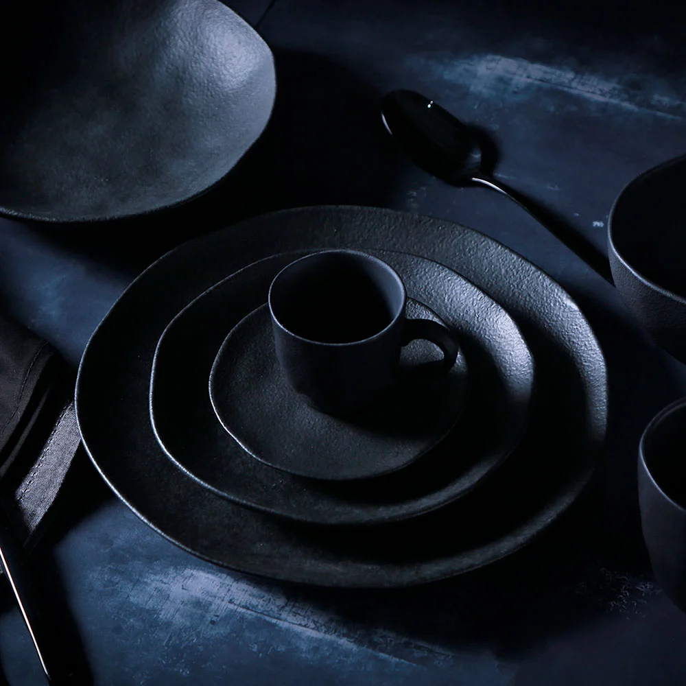 Prato de cerâmica preto com acabamento fosco compondo uma mesa posta elegante, acompanhado de xícara e louças da mesma coleção.