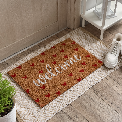 Capacho de porta marrom com estampa “welcome” e corações vermelhos, sobre tapete decorativo na entrada.