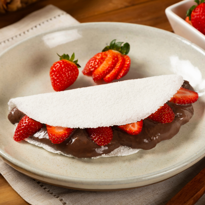 Tapioca recheada com creme de chocolate e morangos fatiados, servida em um prato branco com morangos frescos ao lado.