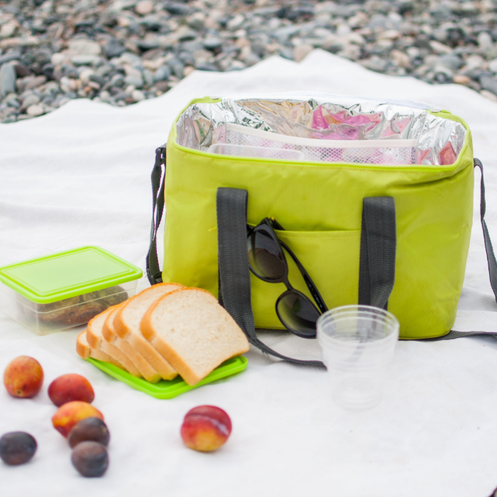Bolsa térmica verde aberta sobre uma toalha branca em uma praia de pedras, acompanhada por fatias de pão, frutas, um pote com tampa verde, óculos escuros e copos plásticos transparentes.
