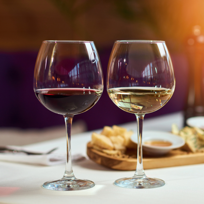 Duas taças de vinho em uma mesa elegante, uma com vinho tinto e outra com vinho branco, acompanhadas por uma tábua de petiscos com queijos e molhos, em um ambiente aconchegante com iluminação suave.
