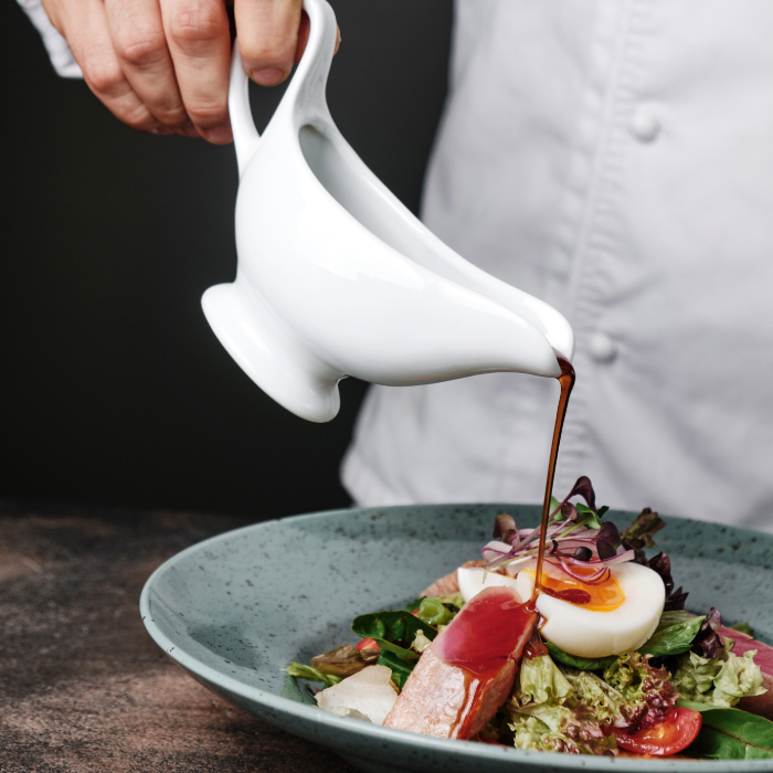 Pessoa despejando molho escuro de uma molheira branca sobre uma salada com folhas verdes, ovo cozido, tomate e filé de peixe, servida em prato fundo verde.