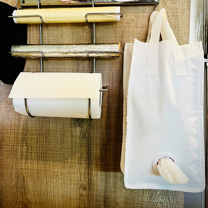 Organizador de cozinha com suporte metálico para papel toalha, papel filme e papel alumínio, ao lado de um porta sacolas plásticas branco pendurado, com uma sacola sendo retirada pela abertura inferior.
