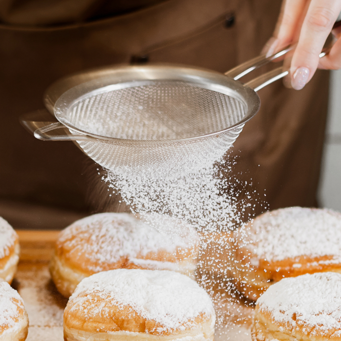 Pessoa polvilhando açúcar de confeiteiro sobre donuts com o uso de uma peneira de inox.