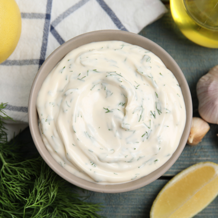 Tigela com molho de alho cremoso com ervas, cercada por ingredientes frescos como limão, alho, endro e uma garrafa de azeite, sobre uma superfície de madeira azul.
