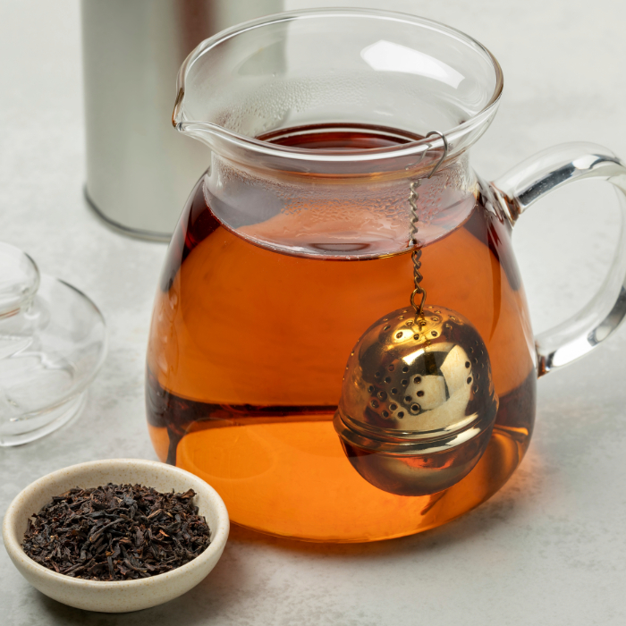 Bule de vidro com chá quente e infusor metálico, acompanhado de um potinho com folhas de chá soltas.