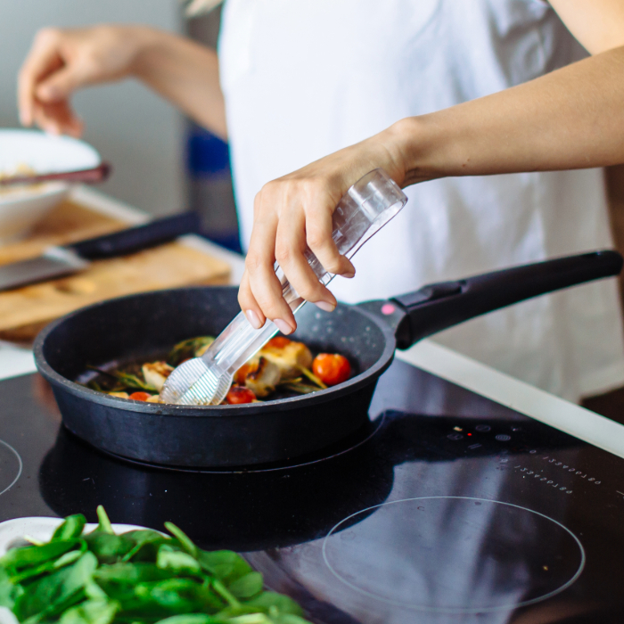 Pessoa temperando alimentos com moedor de pimenta sobre uma frigideira antiaderente em fogão de indução. Na frigideira, há tomates cereja e ervas, e em primeiro plano, folhas verdes frescas sobre a bancada.
