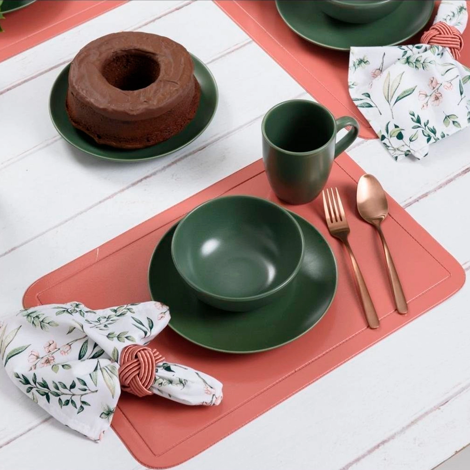 Jogo americano terracota em mesa posta com louças verdes, talheres rosé e guardanapo floral.