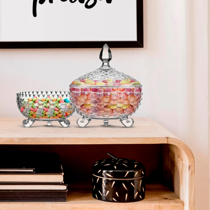 Duas bombonieres de cristal lapidado com tampas, repletas de balas coloridas, dispostas sobre uma prateleira de madeira clara. Ao fundo, um quadro decorativo com uma palavra escrita em preto, complementando a elegância do ambiente.
