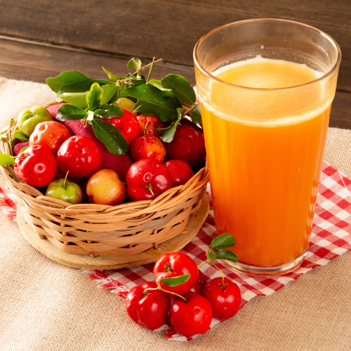 Cesta de vime cheia de acerolas frescas com folhas verdes ao lado de um copo de suco de acerola espumante, sobre um pano xadrez vermelho e branco em uma mesa rústica de madeira.