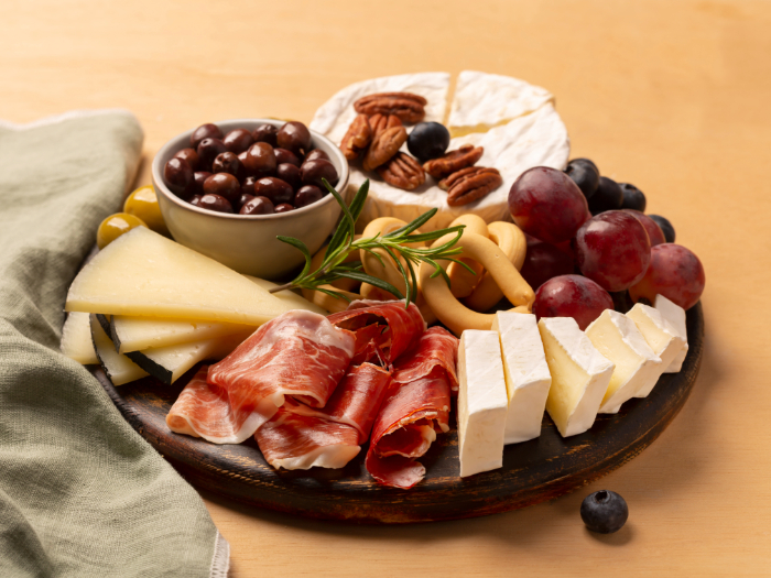 Tábua de frios servida sobre uma superfície de madeira, contendo uma variedade de itens como queijo brie, pedaços de queijo curado, presunto cru, uvas roxas, blueberries, biscoitos salgados, nozes pecan e um ramo de alecrim. Um pequeno recipiente com azeitonas e chocolates cobertos está posicionado ao centro. Ao lado da tábua, há um guardanapo verde, compondo uma apresentação elegante e apetitosa.