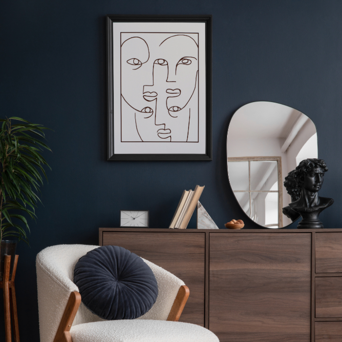 Sala sofisticada com parede azul escura, quadro abstrato de rostos minimalistas, espelho orgânico, aparador de madeira com livros e escultura clássica, além de poltrona bege com almofada redonda azul.