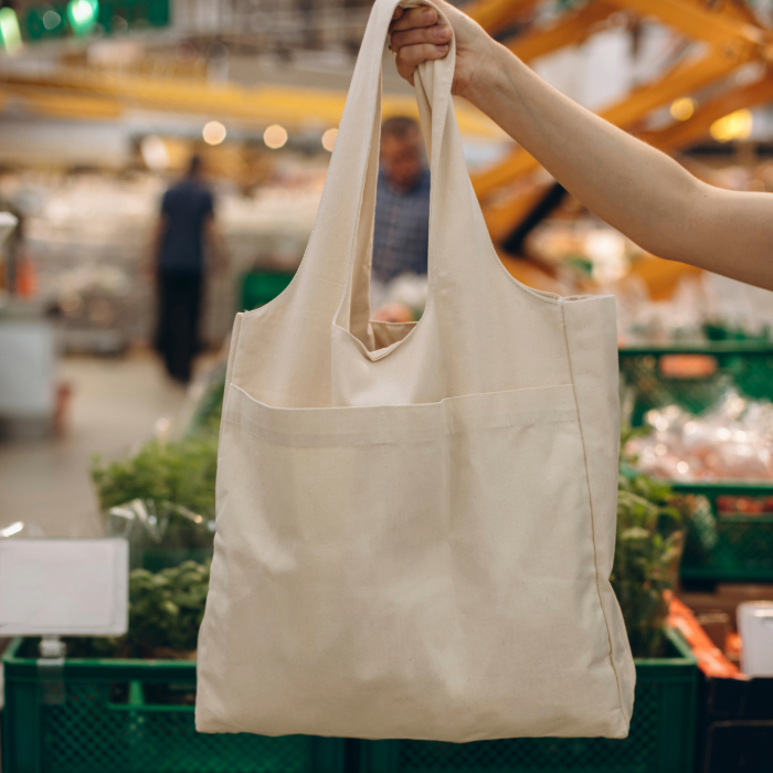 Pessoa segurando uma sacola reutilizável de tecido bege em um mercado, com caixas de vegetais ao fundo.