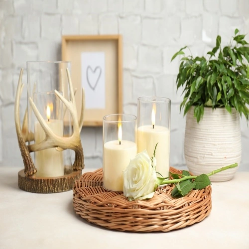 Velas decorativas em porta-velas de vidro e suporte rústico sobre bandeja de vime, acompanhadas de flor branca e planta ornamental em ambiente elegante.