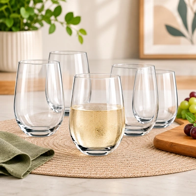 Jogo de copos de vidro transparentes sobre mesa decorada, com um copo servido com bebida clara e frutas ao fundo em ambiente moderno.