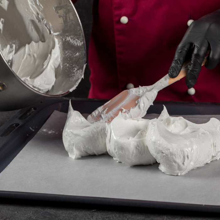 Pessoa usando luvas pretas e jaleco vermelho espalha merengue branco em uma assadeira forrada com papel manteiga, utilizando uma espátula de silicone. Uma tigela com mais merengue aparece parcialmente à esquerda.